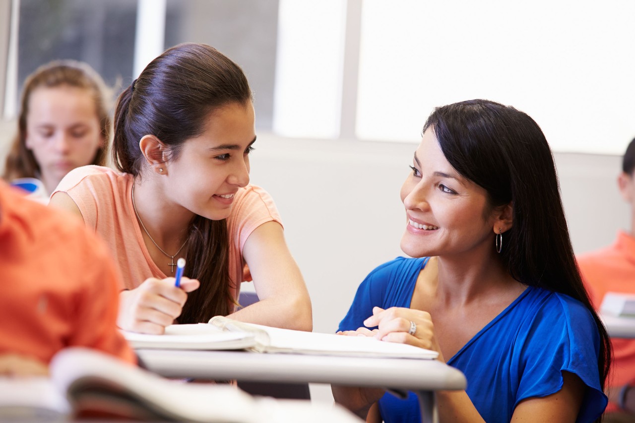 teacher crouching near a student's desk to help, both teacher and student are smiling; fall 2021 return to classroom concept