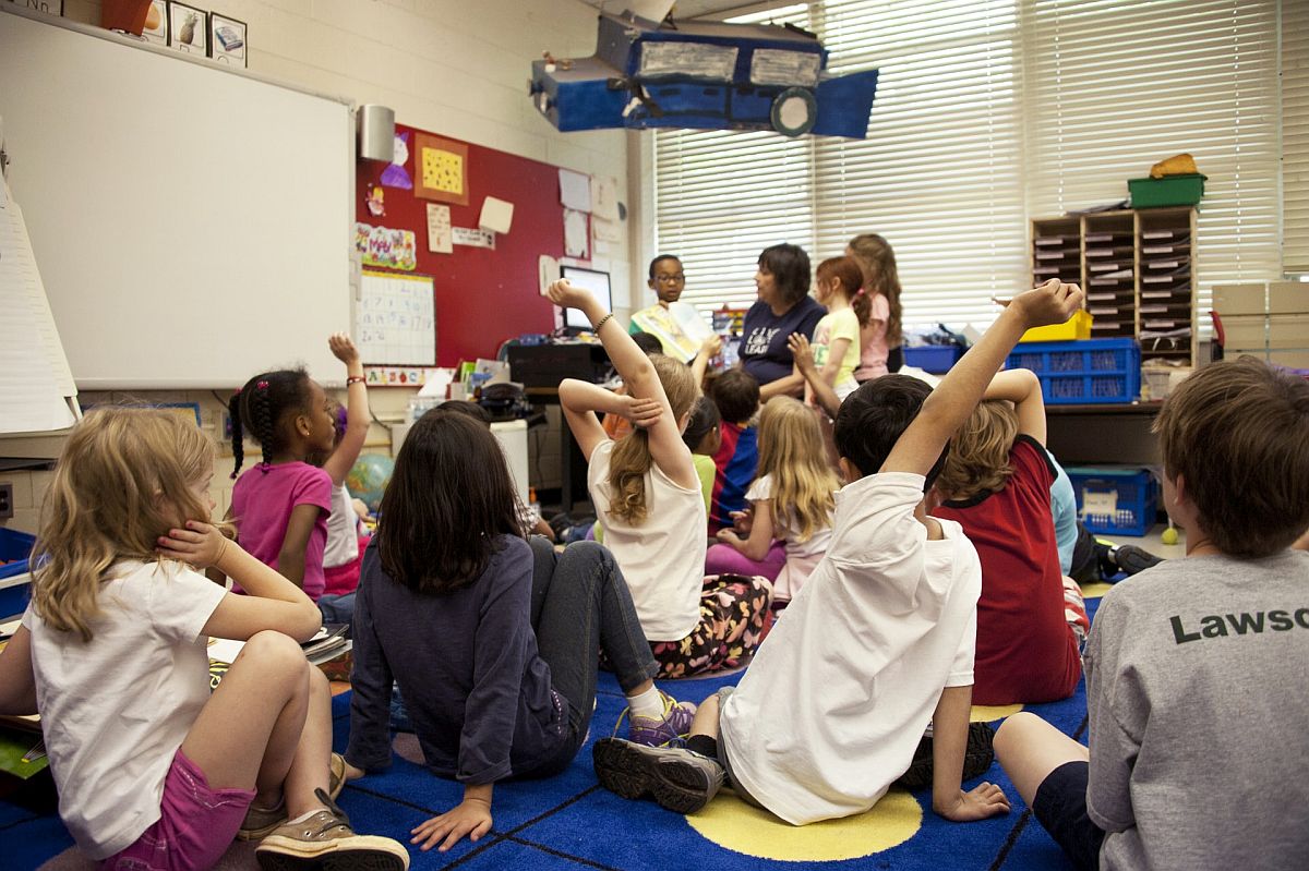 students seated closely together on the floor, raising their hands; concept: classroom changes because of COVID