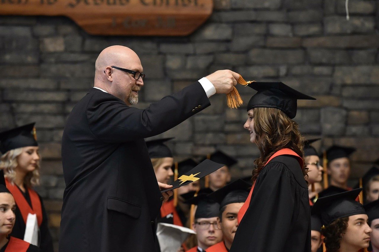 graduation ceremony, principal places mortarboard hat on student; educators guiding students concept