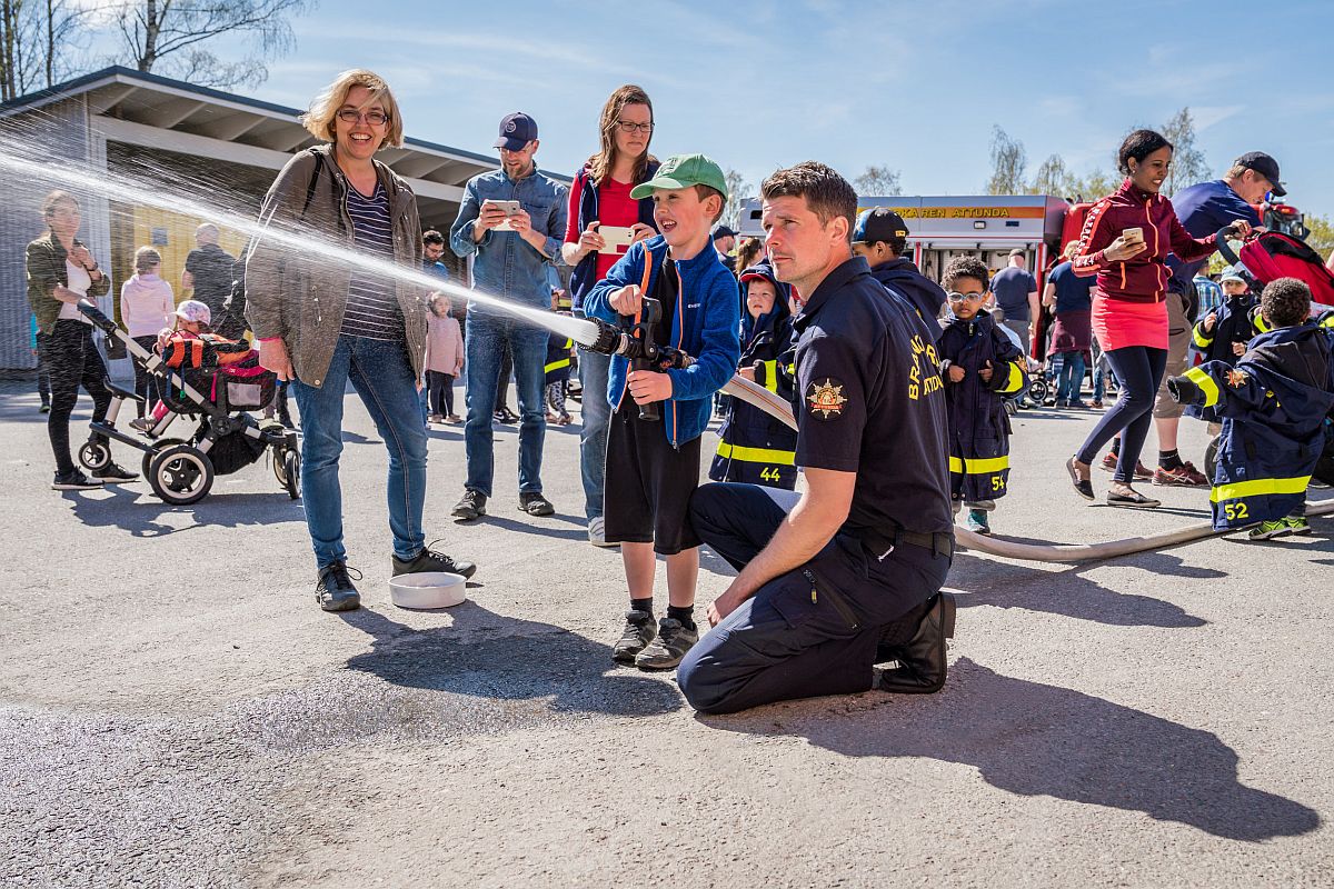 children and parents at a fire station trying a firehose and spraying water, assisted by a fireman; careers lessons plans concept