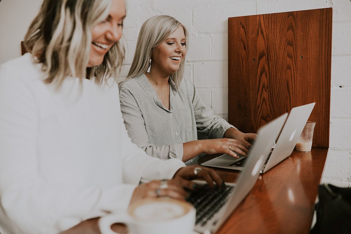 two smiling teachers use laptops beside one another; mental health of teachers concept