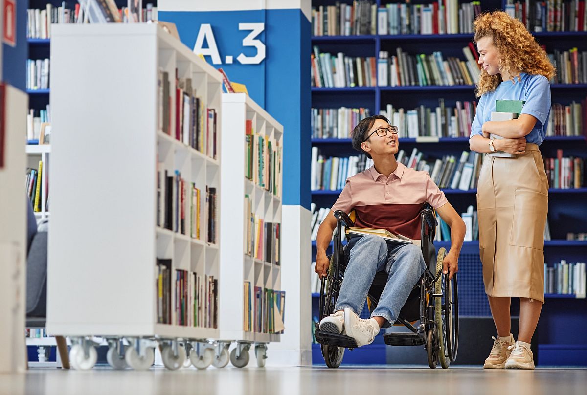 Student in wheelchair looks at young woman holding books in library; paraprofessionals in education concept