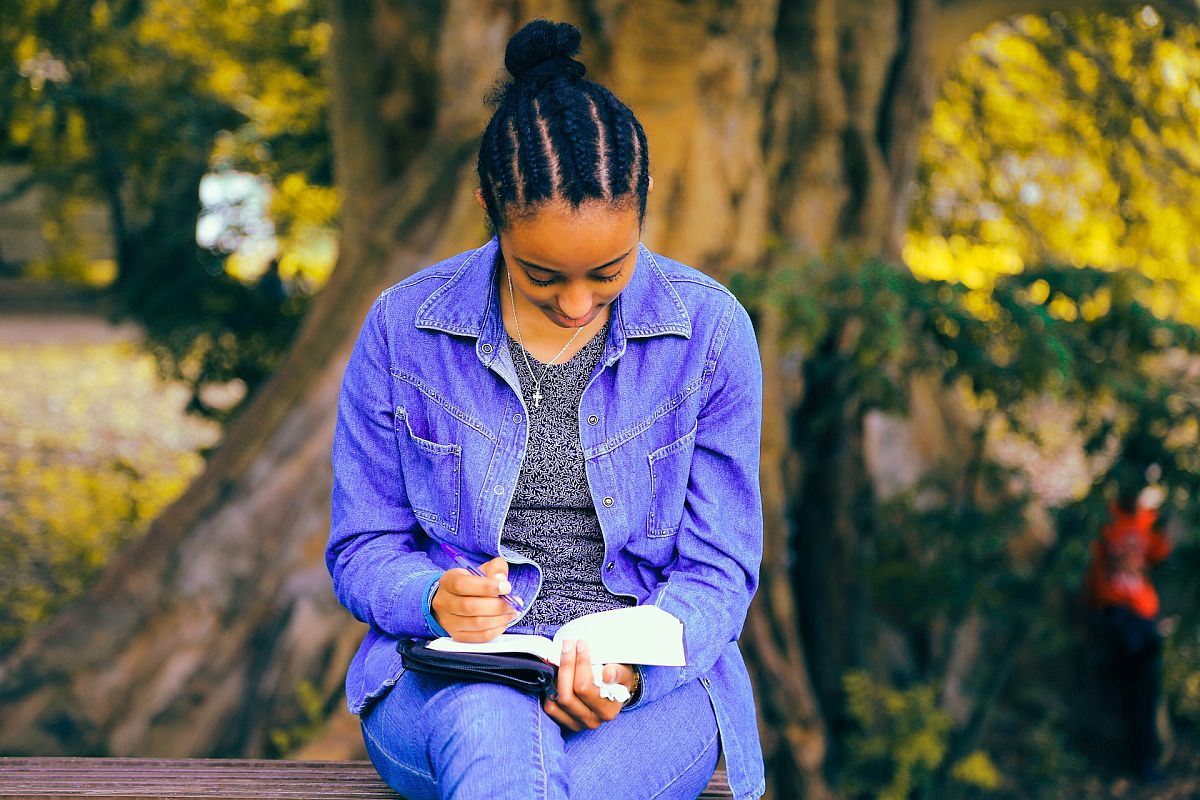 Girl sitting on bench outdoors, reading and making notes in a book; fun literature lesson plans concept