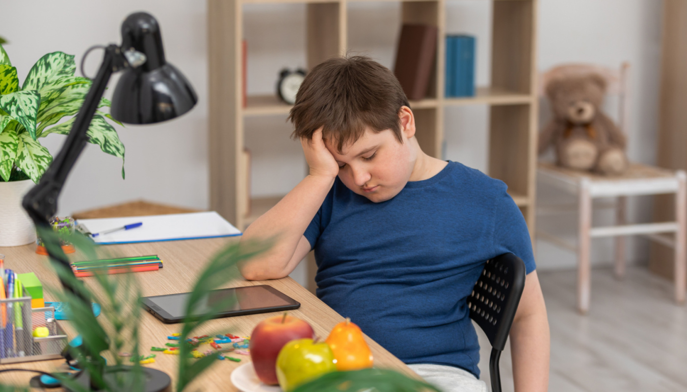 Exhausted boy slumped at his desk, head in his hand; later school start times concept